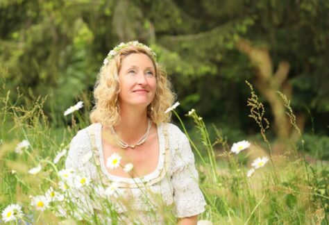 Erzählerin Sonja Fischer von MissFairytale in einem weißen Spitzenkleid sitzt auf einer Wiese im hohen Gras umgeben von Margariten und anderen Gräsern. Auf ihrem blonden gelockten Haar befindet ein Blumenkranz. Sie trägt eine weiße Kette mit Anhänger und blickt lächelnd nach oben. Foto: Max Ciolek