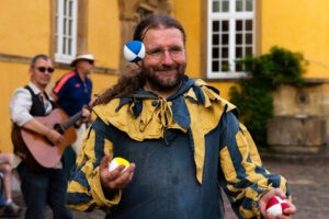 Der Gaukler Olaf to Ossenbrügge steht im Mittelpunkt des Bildes. Er jongliert mit drei Bällen. Er trägt ein blaues Hemd mit gelb-blau gestreiften Kragen und Ärmeln. Im Hintergrun ist verschwommen das Schloß der Universität Osnabrück zu sehen und ein Musiker, der Gitarre spielt. Anlass ist die Kulturnacht XXL zum Jubiläum 350 Jahre Westfälischer Frieden.