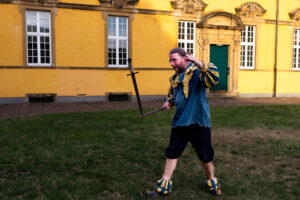Der Gaukler Olaf to Ossenbrügge balanciert ein Schwert auf einer Eisenstange. Er steht vor dem Schloß der Universität Osnabrück. Erträgt ein blaues Hemd mit gelb-blau gestreiften Kragen und Ärmel, eine dunkelblaue Kniebundhose. Anlass ist die Kulturnacht XXL in Osnabrück mit dem Titel Als der Frieden in die Stadt kam.