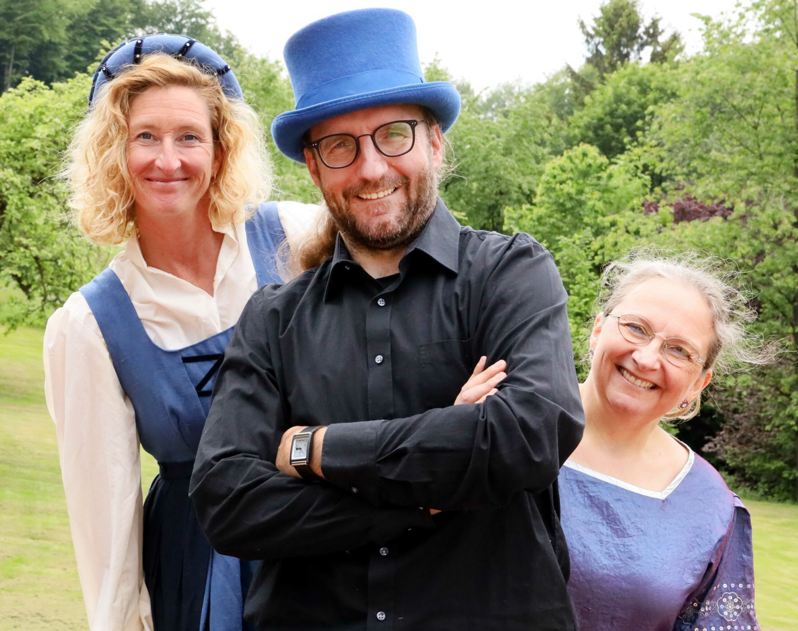 Foto Erzählfinderei, links Erzählerin Soinja Fischer in blau weißem Kostüm, in der Mitte Gaukler Olaf Pieper mit blauem Zylinder, rechts im blauem Kleid Erzählerin Sabine Meyer, Naturhintergrund