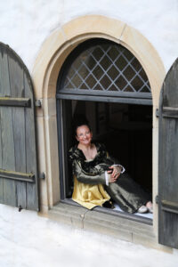 Erzählerin Sabine Meyer im Kostüm. sitzt nach draußen schauend, lächelnd, auf dem Fenstersims eines offenen Flügelfenster