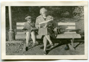 schwarz weiß Foto, Tochter und Mutter sitzen Zeitung lesend auf einer Bank im Sonnenschein, links ein Teil einer Laterne, Naturhintergrund