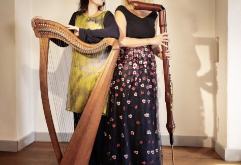 Musikerin Marie Pieck mit Harfe und Erzählerin Sonja Fischer mit Grossbassflöte stehen sich gegenseitig anlächelnd vor einer weißen Wand im Ledenhof.