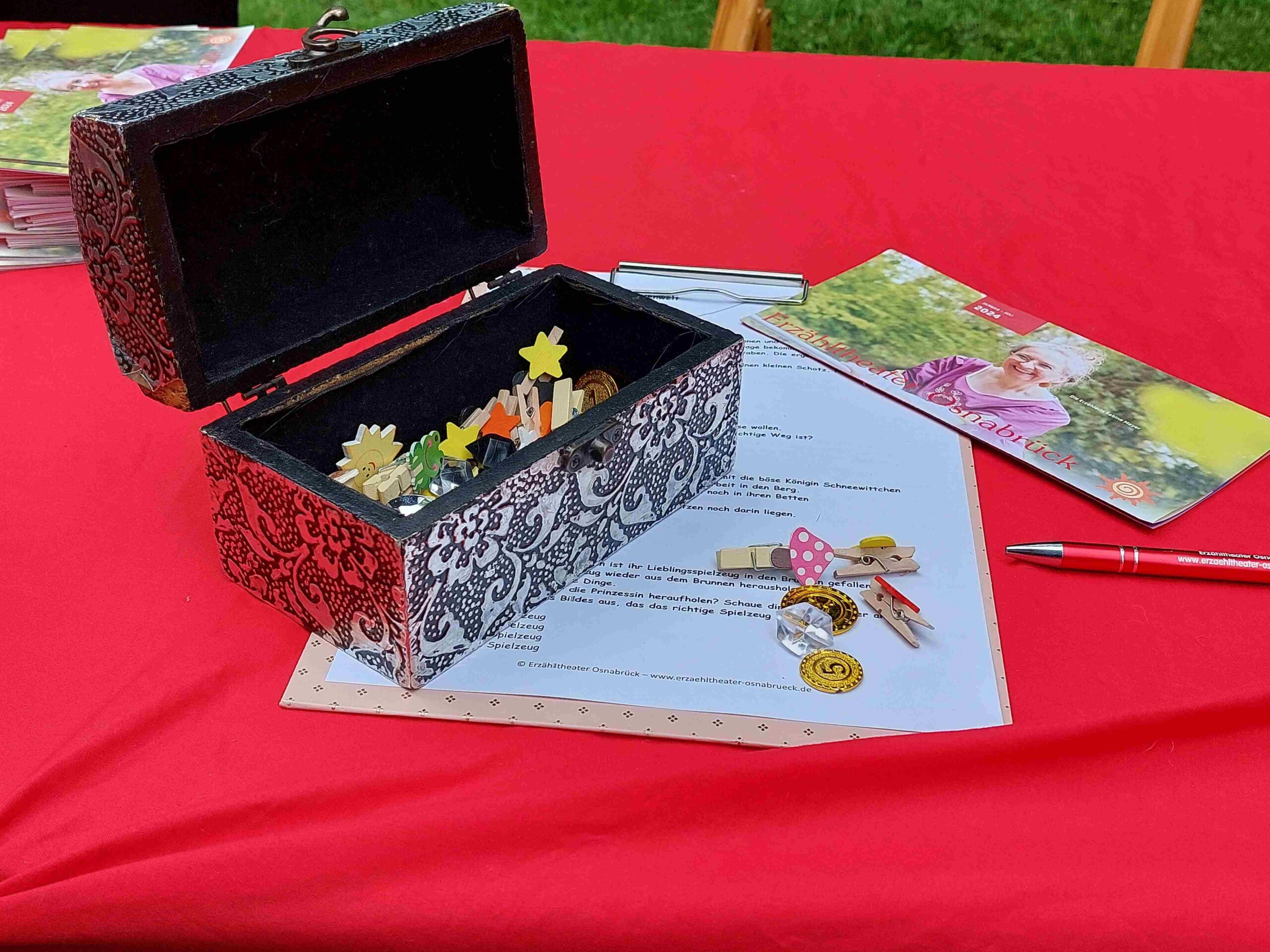 Tisch mit roter Decke, Schatzkiste mit Münzen, Glitzersteinen und verschiedenen Holzklammern mit bunten Sternen, Flyer vom Erzähltheater, Osnabrück, Märchenrallye