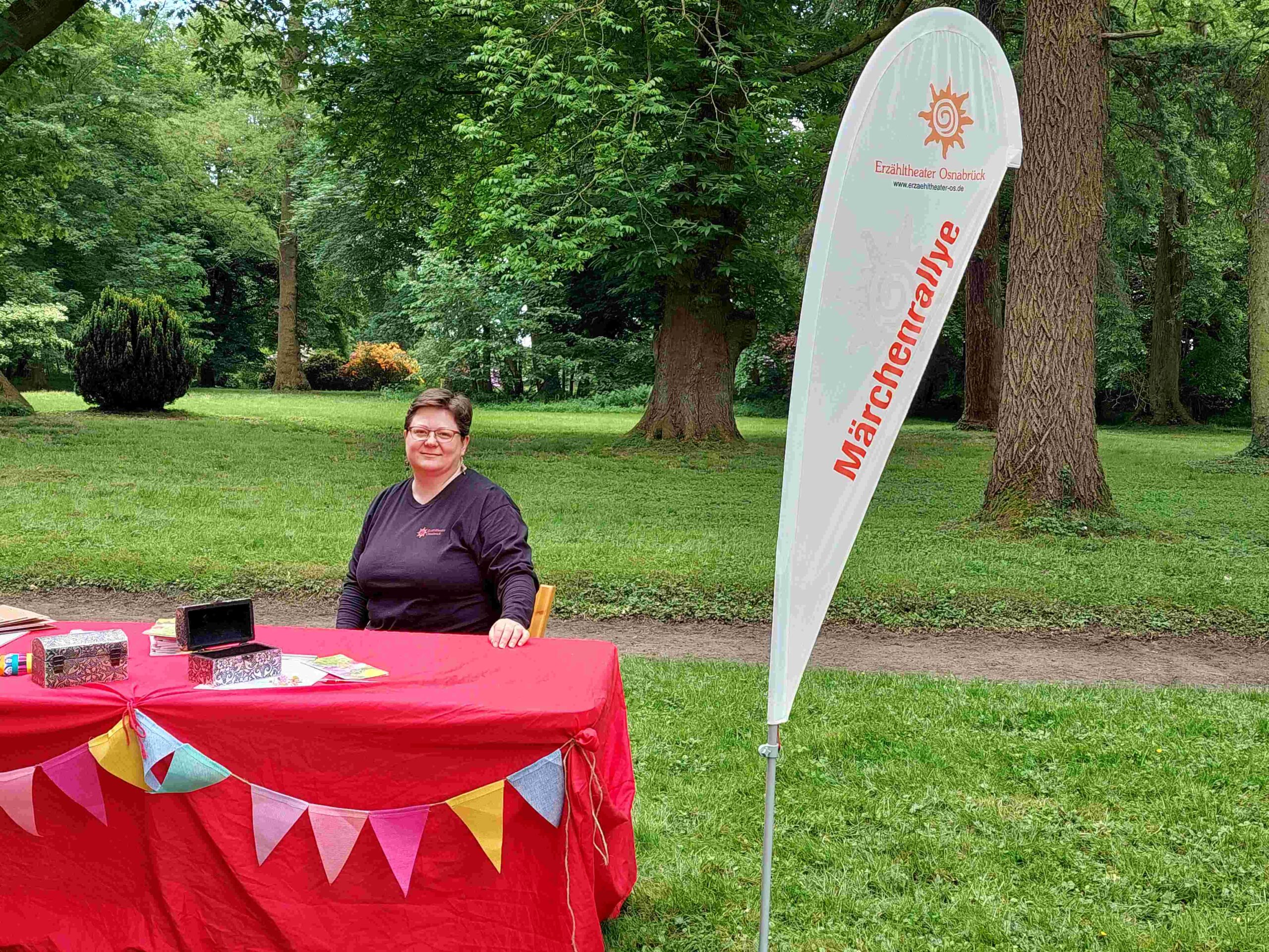 Landschaftsaufnahme. Mitarbeiterin vom Erzähltheater sitzt am roten Tisch mit Wimpeln geschmückt. Daneben die Beach-Flag von der Märchenrallye. Startpunkt der Märchenrallye.