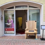 Der Märchenstuhl aus Eichenholz steht auf der geöffneten Tür des Erzähltheaters Osnabrück in der Großen Gildewart 36 in Osnabrück. Neben dem Märchenstuhl steht ein Plakatständer. Auf dem Plakat stehen die Geschichten, die auf dem Märchenstuhl zu hören sind. Links neben der Tür ist ein Plakat mit der Erzählerin Sabine Meyer zu sehen. Sie trägt ein lila Kleid,