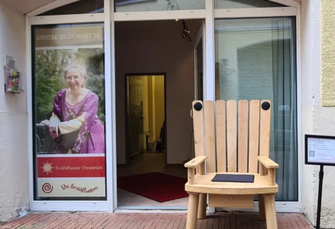 Der Märchenstuhl aus Eichenholz steht auf der geöffneten Tür des Erzähltheaters Osnabrück in der Großen Gildewart 36 in Osnabrück. Neben dem Märchenstuhl steht ein Plakatständer. Auf dem Plakat stehen die Geschichten, die auf dem Märchenstuhl zu hören sind. Links neben der Tür ist ein Plakat mit der Erzählerin Sabine Meyer zu sehen. Sie trägt ein lila Kleid,