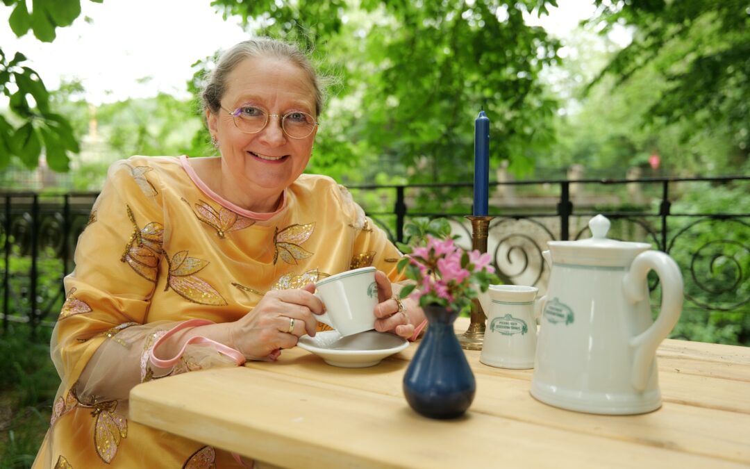 Im Garten an einem Holztisch sitzt die Erzählerin Sabine Meyer mit einer weißen Porzellantasse in der Hand. Vor ihr steht eine alte weiße Porzellankaffeekanne mit dem Logo des Piesberger Gesellschaftshauses.