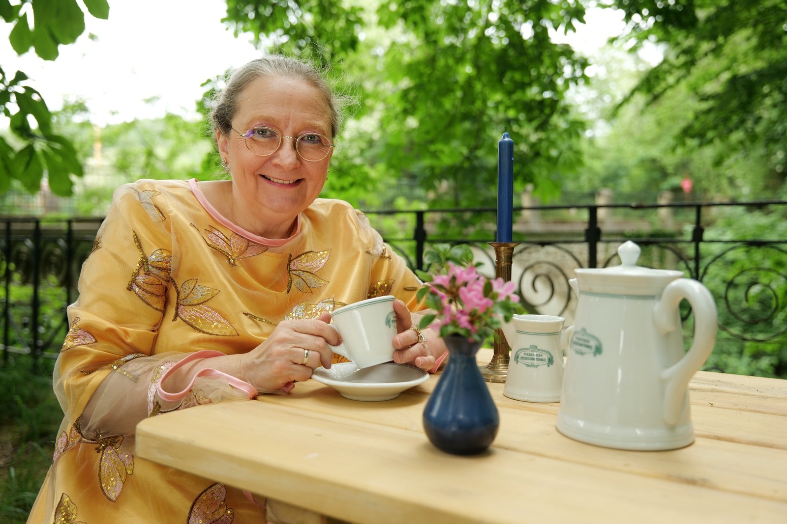 Im Garten an einem Holztisch sitzt die Erzählerin Sabine Meyer mit einer weißen Porzellantasse in der Hand. Vor ihr steht eine alte weiße Porzellankaffeekanne mit dem Logo des Piesberger Gesellschaftshauses.